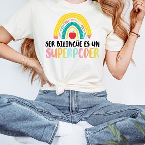 May include: Ivory-colored t-shirt with a rainbow design and the text "SER BILING&Uuml;E ES UN SUPERPODER." The rainbow includes a red apple, pencils, and a ruler. The shirt is tied at the waist and paired with blue jeans.
