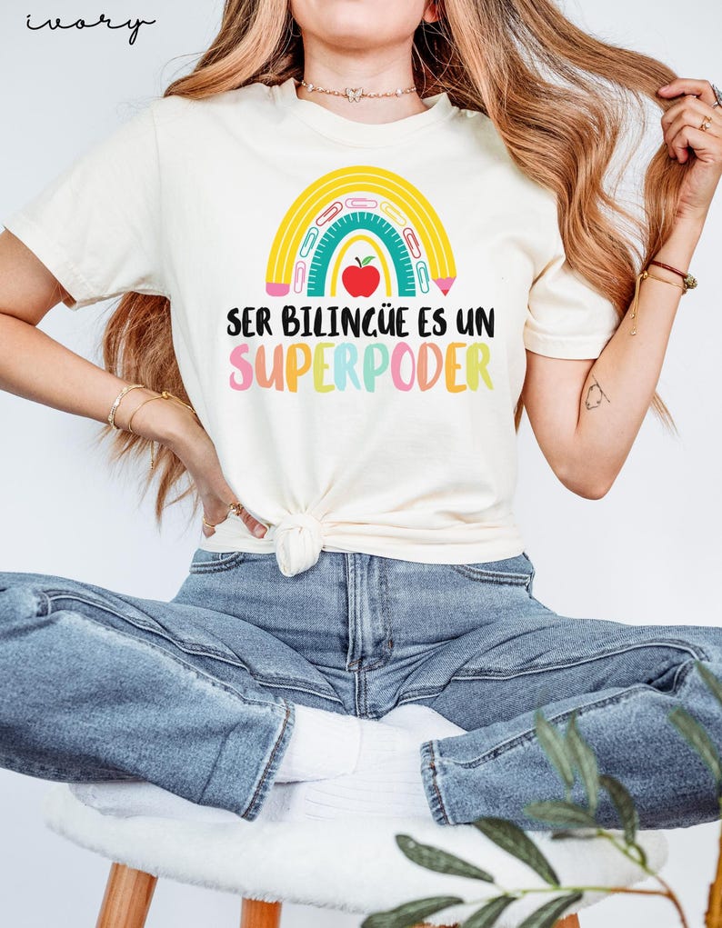 May include: Ivory-colored t-shirt with a rainbow design and the text "SER BILING&Uuml;E ES UN SUPERPODER." The rainbow includes a red apple, pencils, and a ruler. The shirt is tied at the waist and paired with blue jeans.