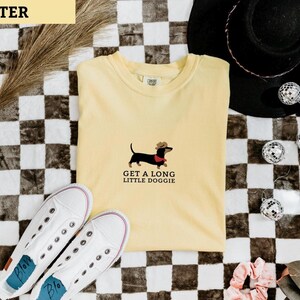 May include: A pale yellow t-shirt featuring a dachshund wearing a cowboy hat and a red scarf, with the text "GET A LONG LITTLE DOGGIE". White trainers and a black hat are also visible in the image.