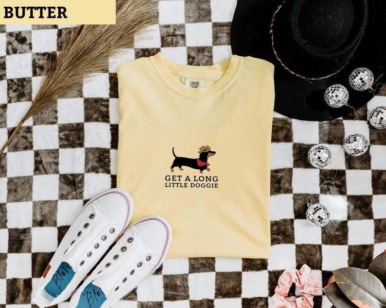 May include: A pale yellow t-shirt featuring a dachshund wearing a cowboy hat and a red scarf, with the text "GET A LONG LITTLE DOGGIE". White trainers and a black hat are also visible in the image.