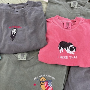 Puede incluir: Una colecci&oacute;n de cinco camisetas de cuello redondo en varios colores, incluyendo gris carb&oacute;n, verde oliva y rosa. Cada camiseta presenta dise&ntilde;os bordados: una m&aacute;scara de fantasma con texto, un perro blanco y negro con el texto I HERD THAT, y un golden retriever sosteniendo una bandera estadounidense con el texto DOG BLESS AMERICA 250TH ANNIVERSARY.