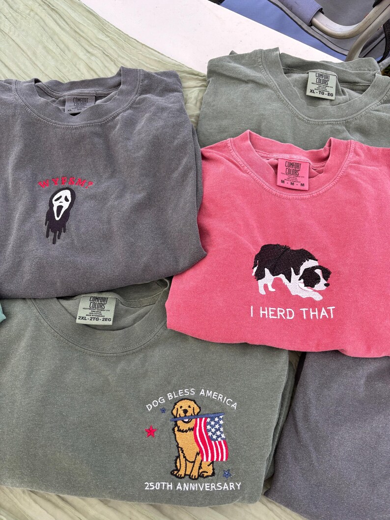 Puede incluir: Una colecci&oacute;n de cinco camisetas de cuello redondo en varios colores, incluyendo gris carb&oacute;n, verde oliva y rosa. Cada camiseta presenta dise&ntilde;os bordados: una m&aacute;scara de fantasma con texto, un perro blanco y negro con el texto I HERD THAT, y un golden retriever sosteniendo una bandera estadounidense con el texto DOG BLESS AMERICA 250TH ANNIVERSARY.