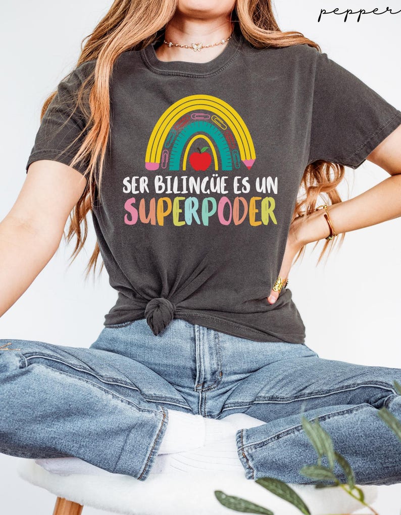 May include: Dark gray t-shirt featuring a rainbow design with a pencil, ruler, and apple. The text "SER BILING&Uuml;E ES UN SUPERPODER" is printed below the rainbow in colorful letters. The shirt is knotted at the waist.