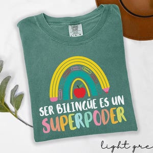 May include: Light green t-shirt with a rainbow design featuring a pencil, paper clips, and a red apple. The text "SER BILING&Uuml;E ES UN SUPERPODER" is printed in colorful letters. The shirt is made of Comfort Colors material.