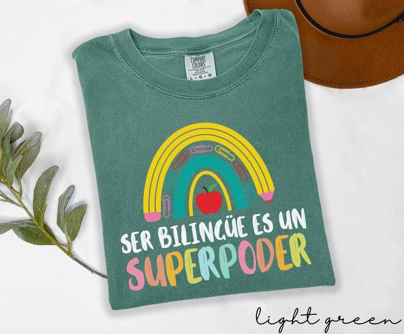 May include: Light green t-shirt with a rainbow design featuring a pencil, paper clips, and a red apple. The text "SER BILING&Uuml;E ES UN SUPERPODER" is printed in colorful letters. The shirt is made of Comfort Colors material.