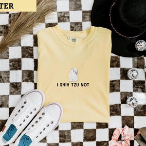 May include: A yellow t-shirt with the text "I Shih Tzu Not" and a small white dog graphic. The shirt is paired with white sneakers and a black hat, all set against a checkered background.