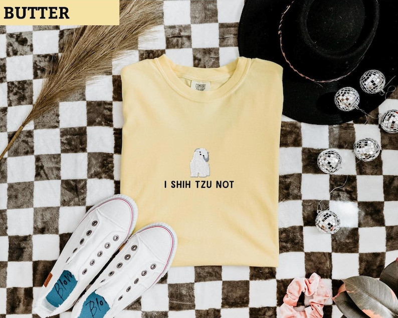 May include: A yellow t-shirt with the text "I Shih Tzu Not" and a small white dog graphic. The shirt is paired with white sneakers and a black hat, all set against a checkered background.