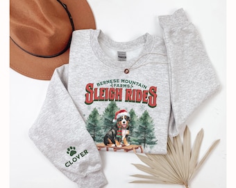 May include: Gray sweatshirt with a Christmas-themed design featuring a Bernese Mountain dog wearing a Santa hat and the text "Bernese Mountain Farms Sleigh Rides". The sweatshirt has a paw print on the sleeve with the word "Clover" underneath.
