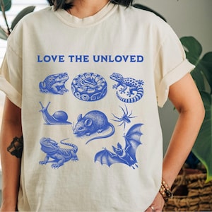 May include: A white t-shirt with the text "LOVE THE UNLOVED" printed in blue. The shirt features a blue illustration of a variety of animals, including a frog, snake, lizard, snail, mouse, spider, and bat.