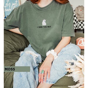 May include: Moss green t-shirt with a small white Shih Tzu graphic and the text "I Shih Tzu Not." The shirt is paired with light blue ripped jeans. A white scrunchie and a large turquoise ring are visible.