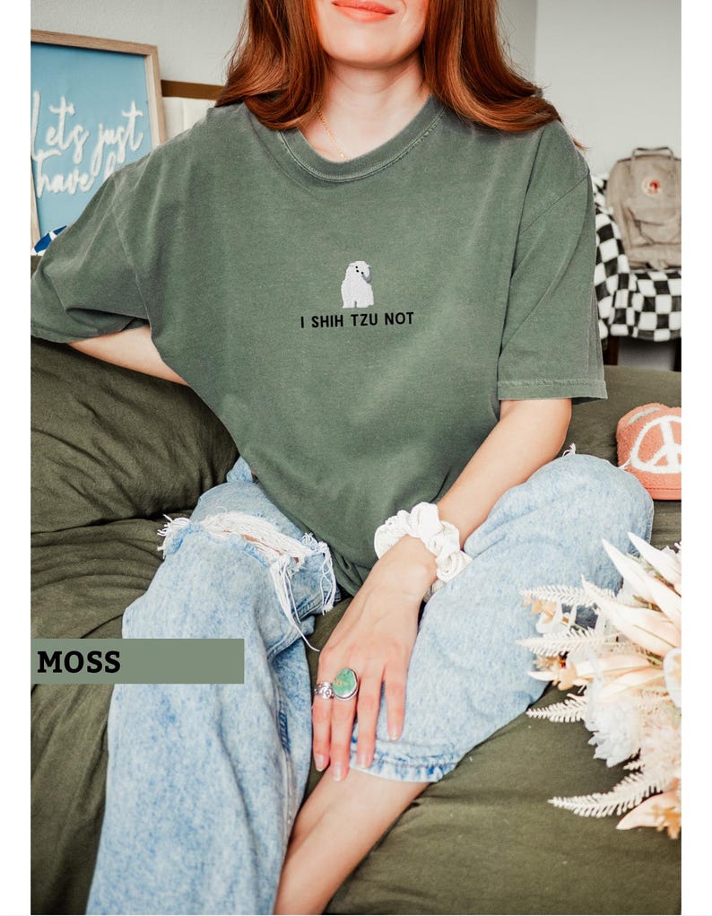 May include: Moss green t-shirt with a small white Shih Tzu graphic and the text "I Shih Tzu Not." The shirt is paired with light blue ripped jeans. A white scrunchie and a large turquoise ring are visible.