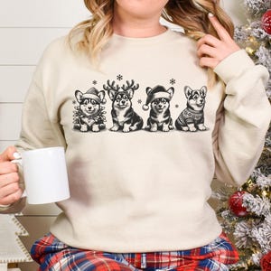 May include: A cream-colored sweatshirt with a festive design featuring four cartoon corgis wearing Santa hats and reindeer antlers. The corgis are arranged in a row and are surrounded by snowflakes.