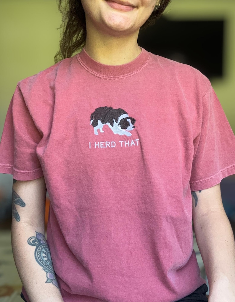 Puede incluir: Una camiseta de manga corta color rosa polvoriento con un bordado de un border collie blanco y negro en posici&oacute;n agachada. Debajo del perro, el texto "I HERD THAT" est&aacute; bordado en letras may&uacute;sculas blancas en el centro del pecho.