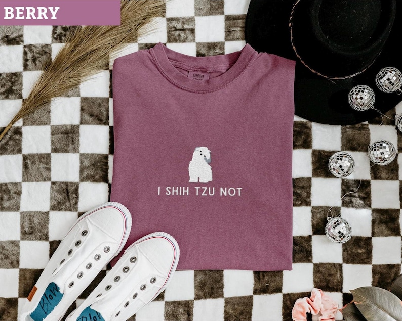 May include: Berry-colored t-shirt with the text "I Shih Tzu Not" and a white Shih Tzu graphic. The shirt is paired with white sneakers, a black hat, and disco ball ornaments. The background has a checkered pattern.