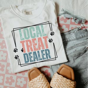 以下が含まれることがあります： 白いTシャツに、カラフルな枠の中に「LOCAL TREAT DEALER」という文字と肉球のプリントがされています。Tシャツはピンクと白の花柄の背景の上に置かれており、茶色のサンダルと青いジーンズが一緒に写っています。