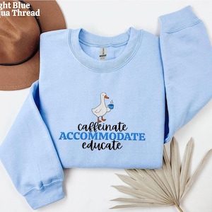Puede incluir: Sudadera azul claro con cuello redondo. La sudadera presenta un diseño bordado de un ganso blanco sosteniendo una taza de café azul, con las palabras "caffeinate, accommodate, educate" en texto azul y negro.