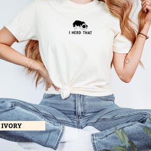 Puede incluir: Una camiseta de manga corta color marfil con un peque&ntilde;o gr&aacute;fico de un perro border collie en blanco y negro sobre el texto negro que dice I HERD THAT. La camiseta tiene cuello redondo y est&aacute; anudada en la parte delantera de la cintura.