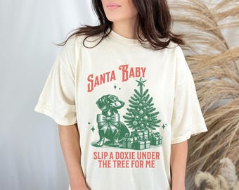 May include: A cream-coloured t-shirt with a green and red graphic print of a dachshund under a Christmas tree. The text on the shirt reads "Santa Baby Slip a Doxie Under the Tree for Me".