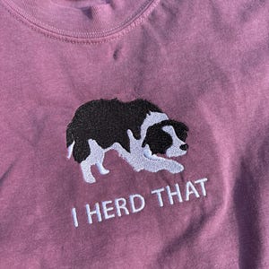 Puede incluir: Una camiseta de color malva con un bordado de un border collie blanco y negro en posici&oacute;n agachada. Debajo del perro, el texto "I HERD THAT" est&aacute; bordado en blanco. La camiseta tiene una etiqueta Comfort Colors en el cuello que indica la talla S.