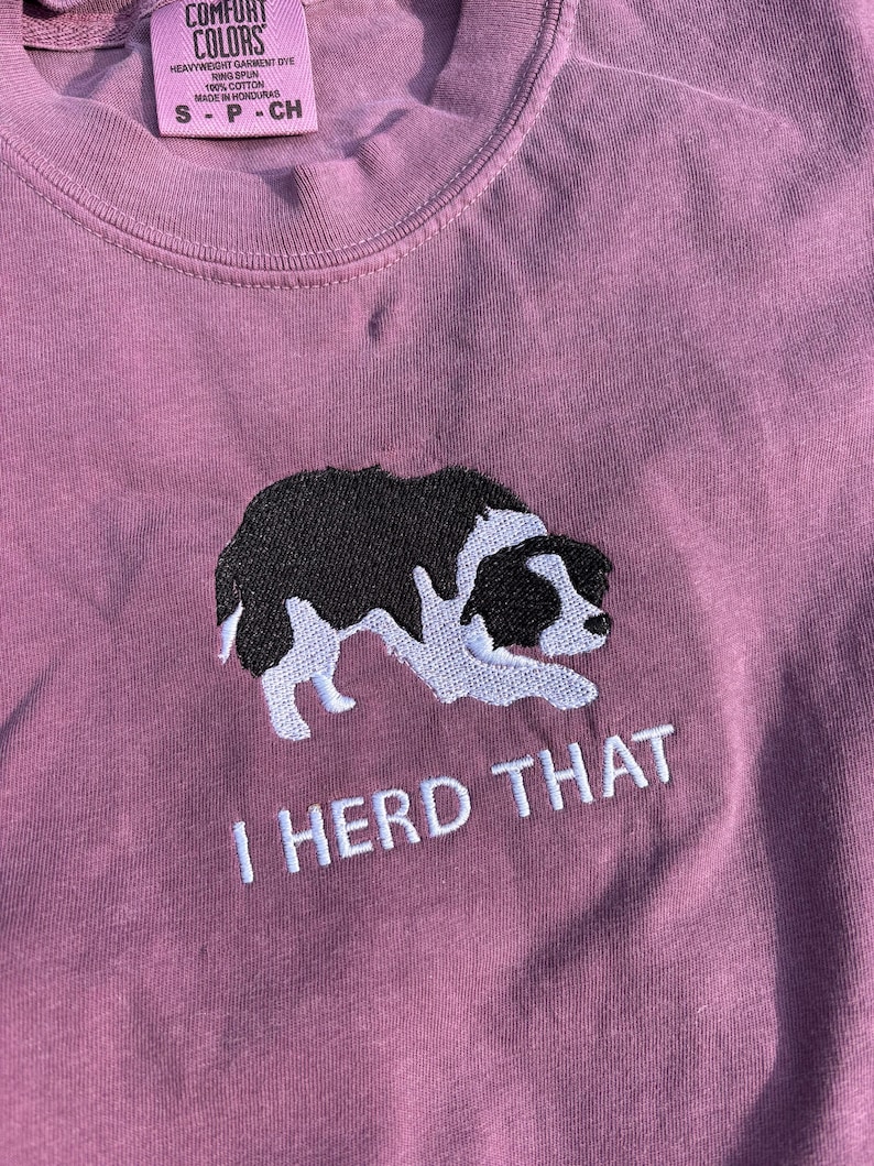 Puede incluir: Una camiseta de color malva con un bordado de un border collie blanco y negro en posici&oacute;n agachada. Debajo del perro, el texto "I HERD THAT" est&aacute; bordado en blanco. La camiseta tiene una etiqueta Comfort Colors en el cuello que indica la talla S.