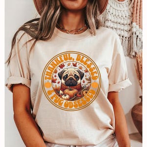 May include: A beige t-shirt with a graphic print of a pug surrounded by fall leaves and pumpkins. The text on the shirt reads "Thankful, Blessed & Pug Obsessed".