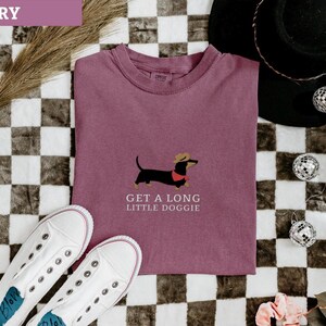 May include: Berry-coloured t-shirt featuring a dachshund graphic wearing a hat and a red collar. The text "GET A LONG LITTLE DOGGIE" is below the dog. White trainers are at the bottom, and a black hat and disco balls are in the background.