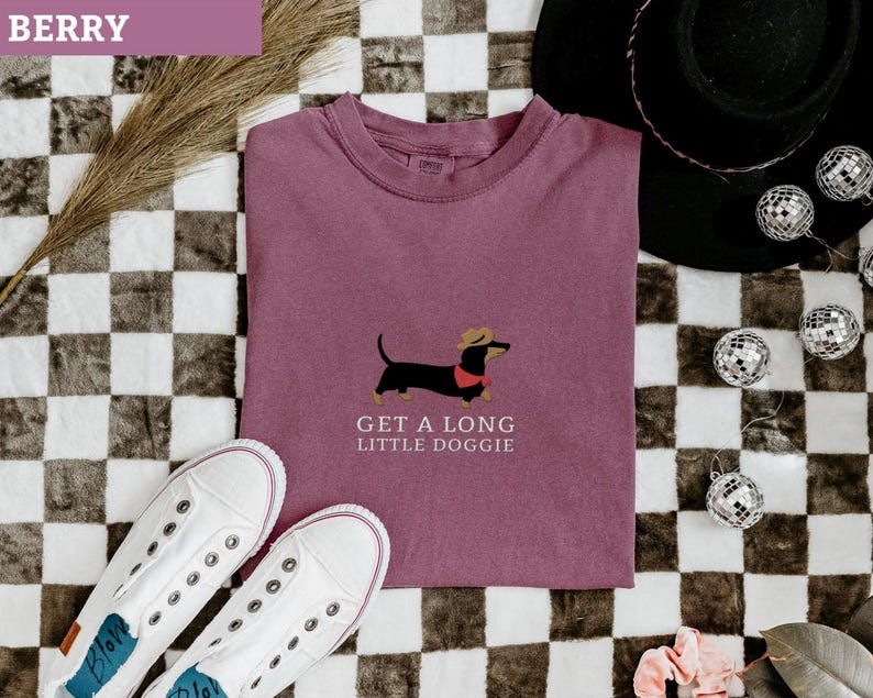 May include: Berry-coloured t-shirt featuring a dachshund graphic wearing a hat and a red collar. The text "GET A LONG LITTLE DOGGIE" is below the dog. White trainers are at the bottom, and a black hat and disco balls are in the background.
