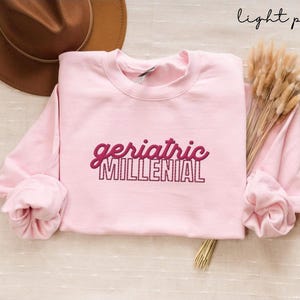 Puede incluir: Sudadera rosa claro con el texto "geriatric MILLENIAL" en un diseño de letras cursivas y en bloque. Un sombrero marrón y flores secas son visibles en el fondo. Las palabras "light pink" están escritas en cursiva en la esquina superior derecha.