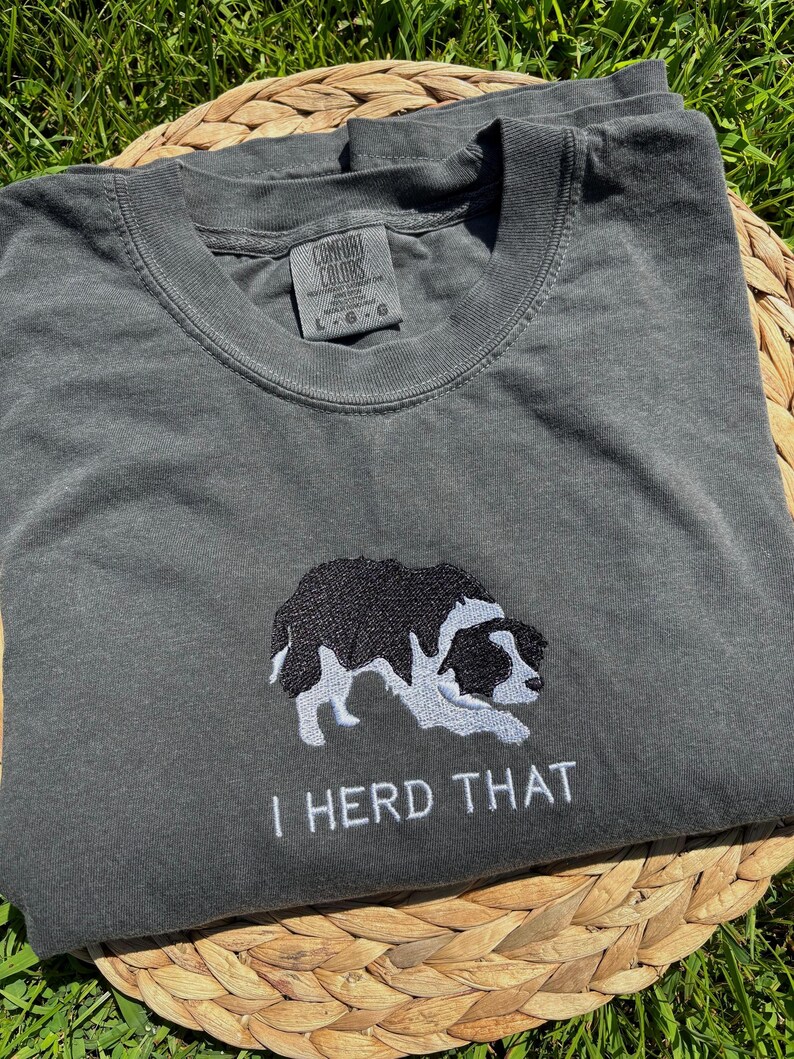 Puede incluir: Una camiseta de cuello redondo gris carb&oacute;n con un bordado de un border collie blanco y negro en posici&oacute;n agachada. Debajo del perro, el texto "I HERD THAT" est&aacute; bordado en hilo blanco. La camiseta tiene una etiqueta de marca Comfort Colors.