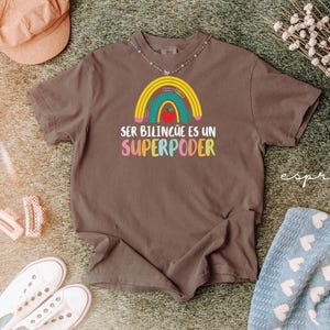May include: A brown t-shirt with a colorful rainbow design and the text "SER BILING&Uuml;E ES UN SUPERPODER". The shirt is surrounded by a peach-colored hat, white sneakers, and a blue patterned blanket.