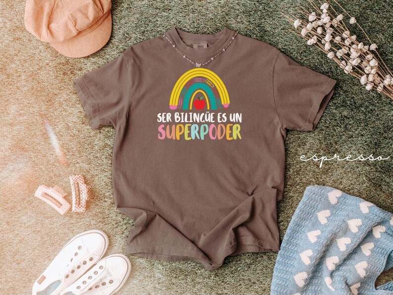 May include: A brown t-shirt with a colorful rainbow design and the text "SER BILING&Uuml;E ES UN SUPERPODER". The shirt is surrounded by a peach-colored hat, white sneakers, and a blue patterned blanket.