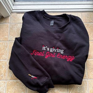 Peut inclure: Un sweat-shirt noir à col rond avec une broderie blanche et rouge sur la poitrine indiquant "it's giving Final Girl Energy". La manche gauche présente un petit motif brodé représentant un couteau argenté avec une touche rouge sang.