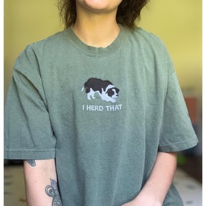 Puede incluir: Una camiseta de manga corta color verde oliva con un bordado de un border collie blanco y negro en posici&oacute;n agachada. Debajo del perro, el texto "I HERD THAT" est&aacute; bordado en letras may&uacute;sculas blancas en la zona del pecho de la prenda.