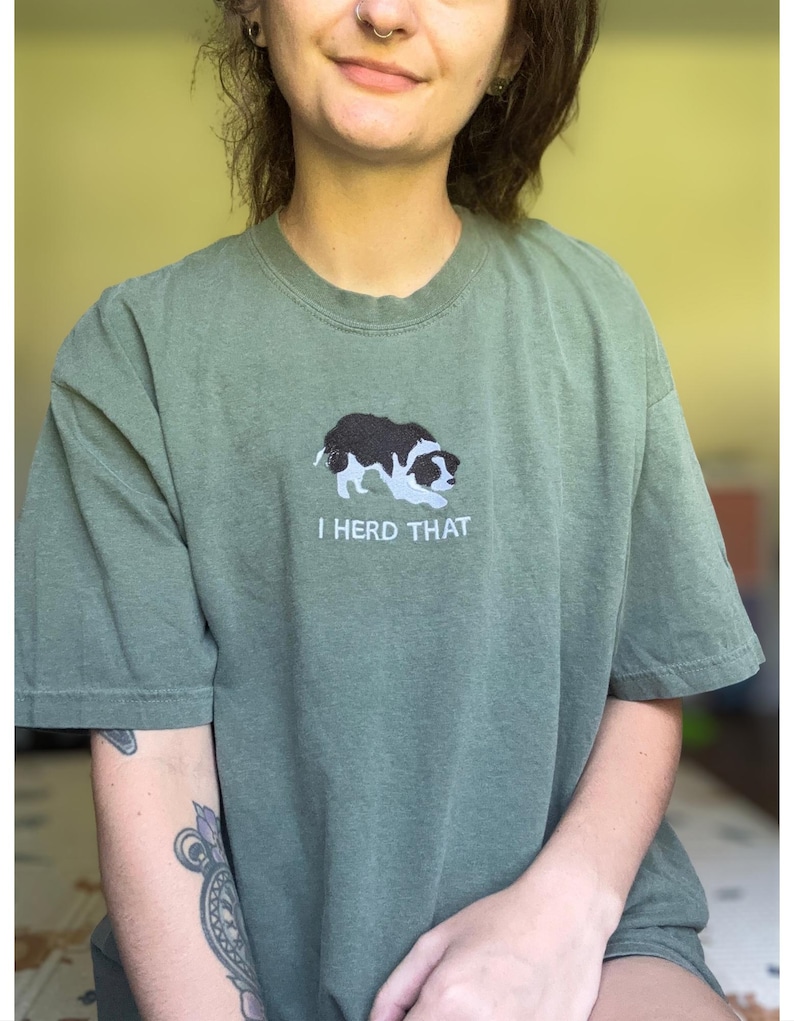 Puede incluir: Una camiseta de manga corta color verde oliva con un bordado de un border collie blanco y negro en posici&oacute;n agachada. Debajo del perro, el texto "I HERD THAT" est&aacute; bordado en letras may&uacute;sculas blancas en la zona del pecho de la prenda.