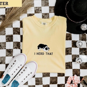 Puede incluir: Una camiseta de manga corta color amarillo mantequilla con una ilustraci&oacute;n de un border collie en blanco y negro centrada en el pecho y el texto en negro I HERD THAT impreso justo debajo del gr&aacute;fico del perro en una fuente sencilla.