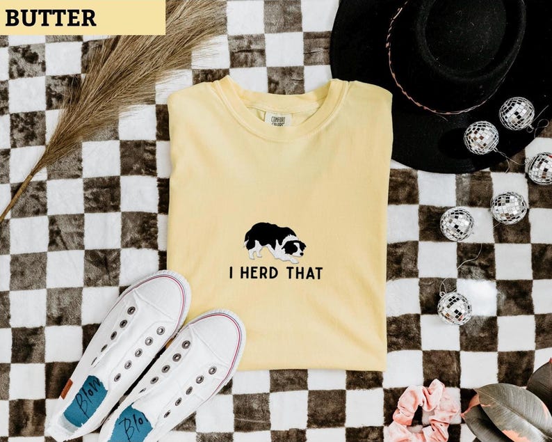 Puede incluir: Una camiseta de manga corta color amarillo mantequilla con una ilustraci&oacute;n de un border collie en blanco y negro centrada en el pecho y el texto en negro I HERD THAT impreso justo debajo del gr&aacute;fico del perro en una fuente sencilla.