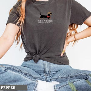 May include: Dark grey t-shirt with a dachshund illustration wearing a hat and red collar. The text "GET A LONG LITTLE DOGGIE" is printed below the dog. The shirt is a casual style.