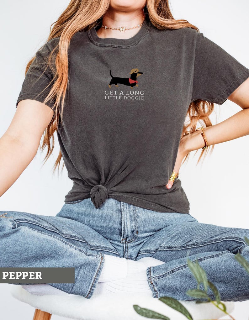May include: Dark grey t-shirt with a dachshund illustration wearing a hat and red collar. The text "GET A LONG LITTLE DOGGIE" is printed below the dog. The shirt is a casual style.