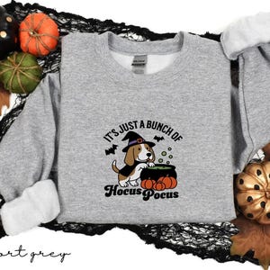 Puede incluir: Sudadera gris deportiva con un diseño de Halloween. El diseño presenta un beagle con un sombrero de bruja, un caldero y calabazas, con el texto "It's just a bunch of Hocus Pocus". Calabazas y hojas decorativas rodean la sudadera.