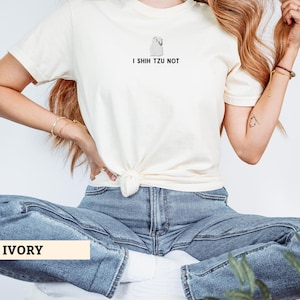May include: Ivory-colored t-shirt with a graphic of a Shih Tzu dog and the text "I Shih Tzu Not." The shirt is knotted at the waist and paired with blue jeans. The word "IVORY" is displayed in a rectangular box.
