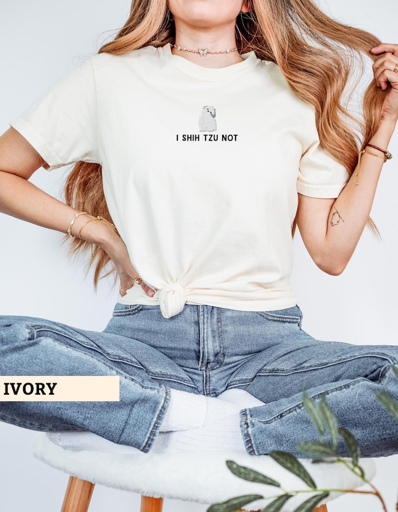 May include: Ivory-colored t-shirt with a graphic of a Shih Tzu dog and the text "I Shih Tzu Not." The shirt is knotted at the waist and paired with blue jeans. The word "IVORY" is displayed in a rectangular box.