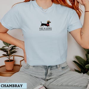 May include: Pale blue t-shirt featuring a dachshund illustration wearing a cowboy hat and a red scarf. The text "GET A LONG LITTLE DOGGIE" is printed below the dog. The shirt is made of a soft material.