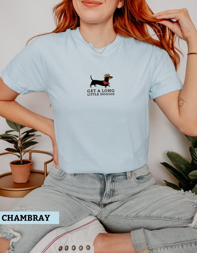 May include: Pale blue t-shirt featuring a dachshund illustration wearing a cowboy hat and a red scarf. The text "GET A LONG LITTLE DOGGIE" is printed below the dog. The shirt is made of a soft material.