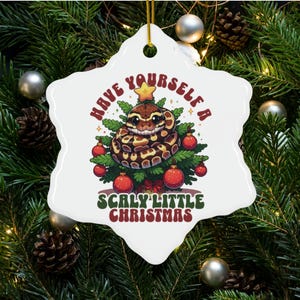 May include: White ceramic snowflake ornament with a cartoon snake wrapped around a Christmas tree. The ornament has the text "Have yourself a scaly little Christmas".