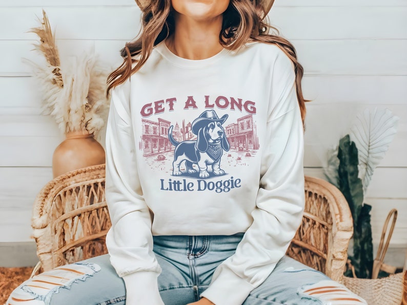 May include: A white long-sleeved sweatshirt with a graphic print of a cartoon basset hound wearing a cowboy hat and bandana. The text "Get a Long Little Doggie" is printed below the image.