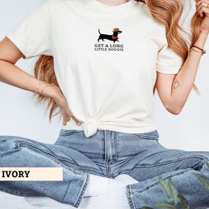 May include: Ivory t-shirt with a dachshund wearing a cowboy hat and a red scarf. The text "GET A LONG LITTLE DOGGIE" is printed below the dog illustration. The shirt is tied at the waist.