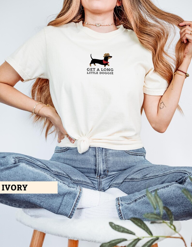 May include: Ivory t-shirt with a dachshund wearing a cowboy hat and a red scarf. The text "GET A LONG LITTLE DOGGIE" is printed below the dog illustration. The shirt is tied at the waist.
