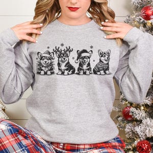 May include: A gray sweatshirt with a festive design featuring five cartoon corgis wearing Santa hats and reindeer antlers. The corgis are arranged in a row and are all smiling. The sweatshirt has a snowflake pattern.