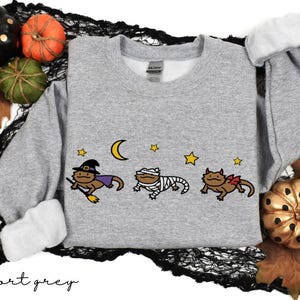 May include: A light gray sweatshirt with a Halloween-themed graphic. The graphic features three cartoon lizards dressed as a witch, mummy, and a lizard with a bow. A crescent moon and stars are above the lizards. Decorative pumpkins and leaves surround the sweatshirt.