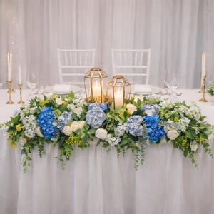 Könnte beinhalten: Eine festlich gedeckte Tafel mit weißer Tischdecke, Stühlen und einem Blumengesteck. Der Mittelpunkt besteht aus blauen Hortensien, weißen Rosen und Grün, mit goldenen Laternen und Kerzen. Elegant und romantisch.